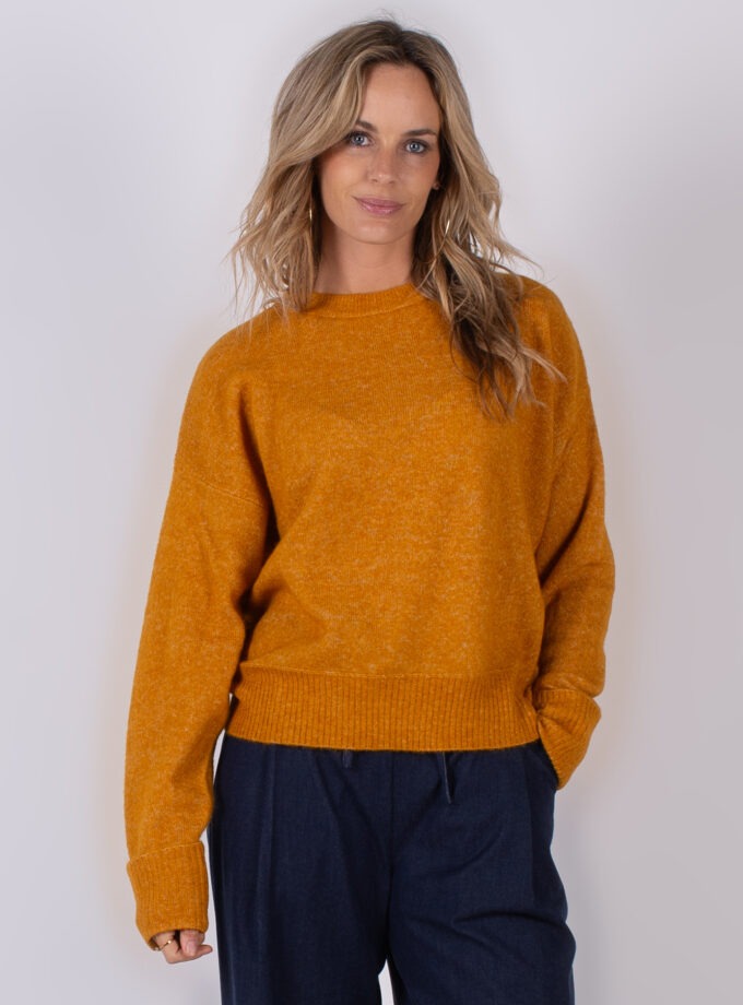 Vrouw poseert in een okergele gebreide trui en donkerblauwe broek uit de Studio AW25-26 collectie.