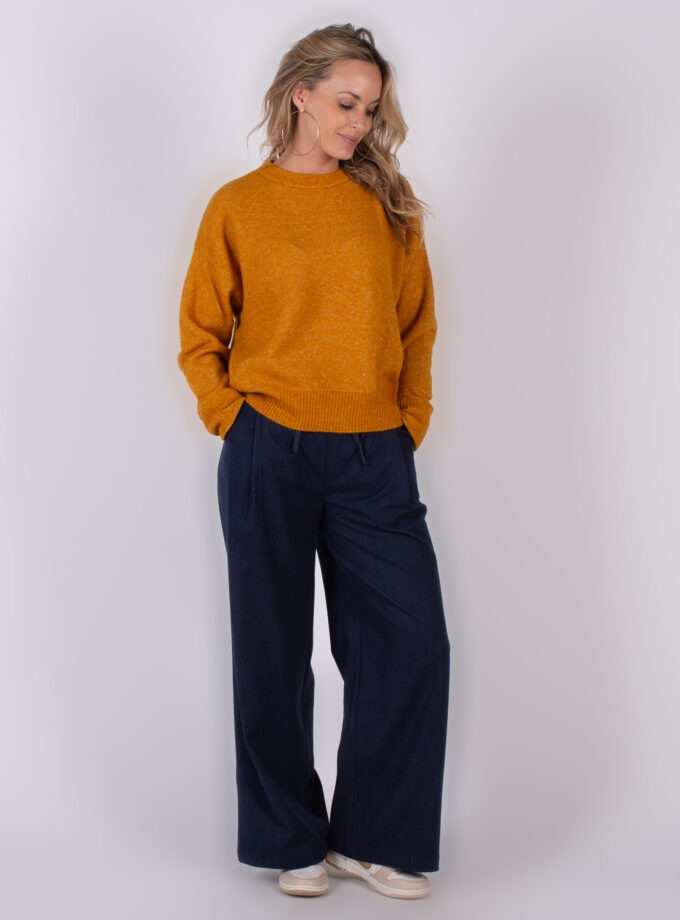 Vrouw draagt een okergele trui en wijde donkerblauwe broek met sneakers in een studio-opname.