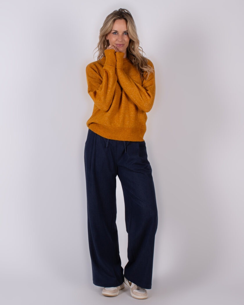 Vrouw poseert in een okergele trui en wijde donkerblauwe broek uit de Studio_AW25-26#2-209 collectie.