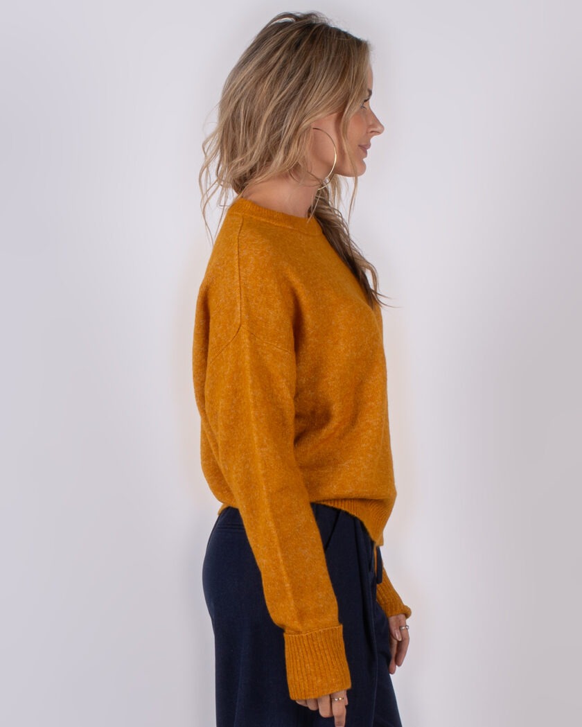 Vrouw in profiel poseert in okergele trui en donkerblauwe broek tegen een neutrale achtergrond.