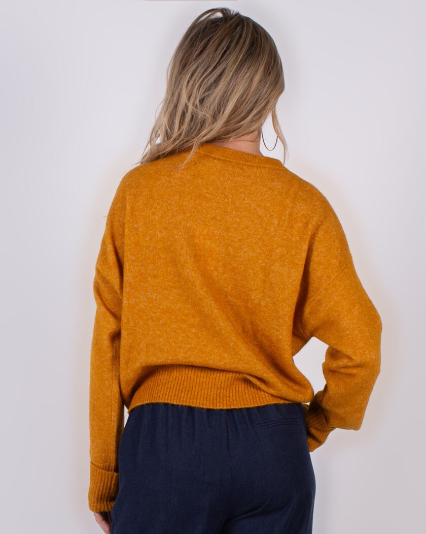 Vrouw van achteren in een mosterdgele trui en donkerblauwe broek uit de Studio AW25-26 collectie.