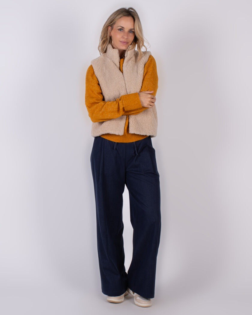 Vrouw in een Studio_AW25-26#2-213 outfit met beige teddy bodywarmer, okergele trui en donkerblauwe wijde broek.