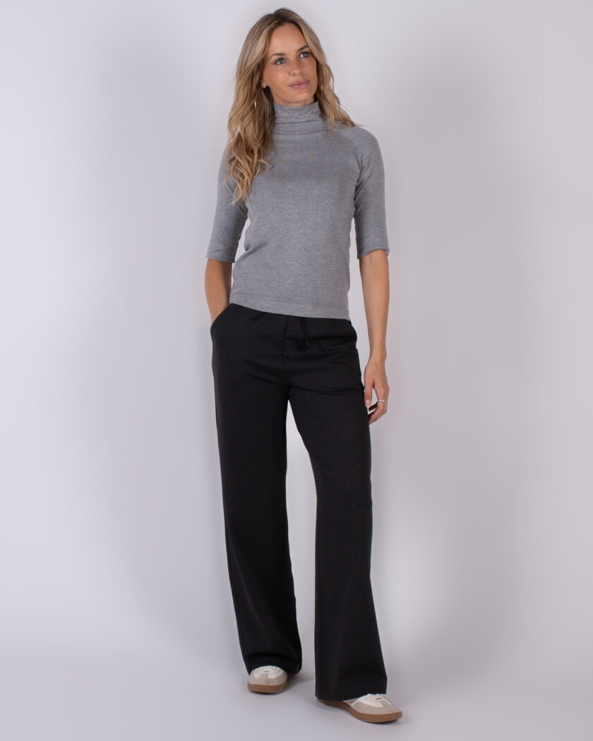 Vrouw draagt een grijze coltrui met halve mouwen en zwarte, wijde broek uit de Studio AW25-26 collectie.