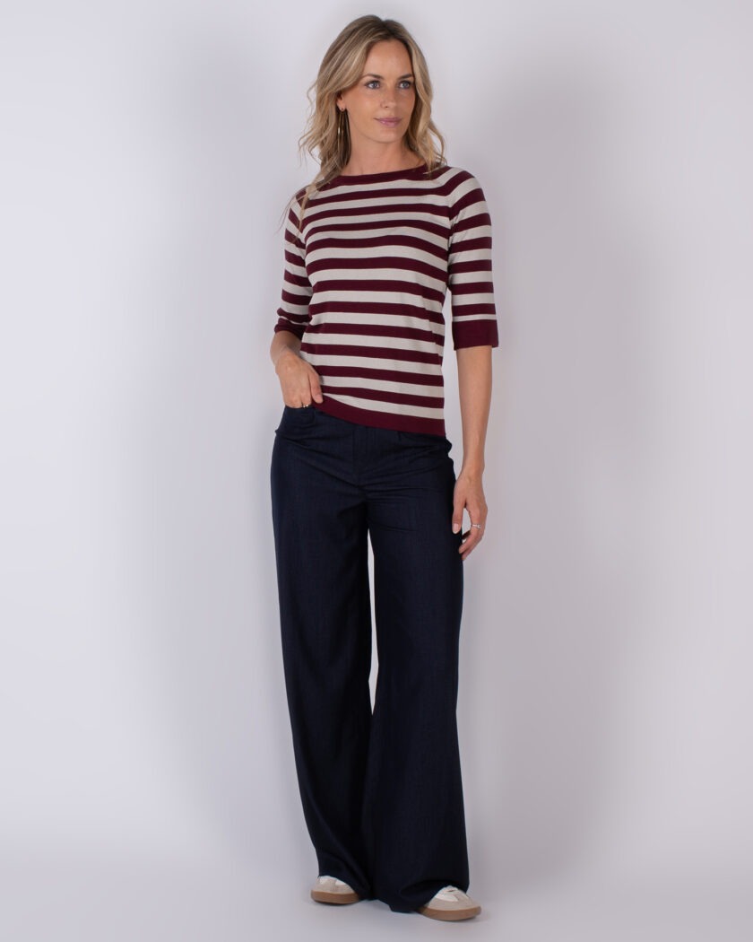 Vrouw in een bordeaux-rood met beige gestreepte top en donkerblauwe wijde broek poseert tegen een neutrale achtergrond.