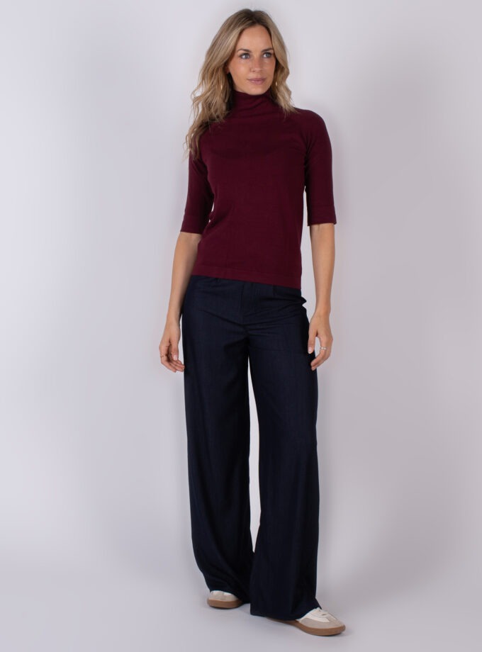 Vrouw draagt een bordeauxrode coltrui met halve mouwen en donkerblauwe wide leg broek uit de Studio AW25-26 collectie.