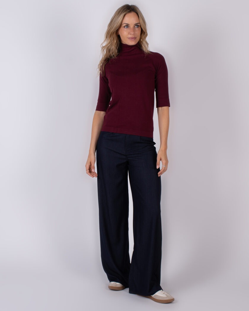 Vrouw draagt een bordeauxrode coltrui met halve mouwen en donkerblauwe wide leg broek uit de Studio AW25-26 collectie.