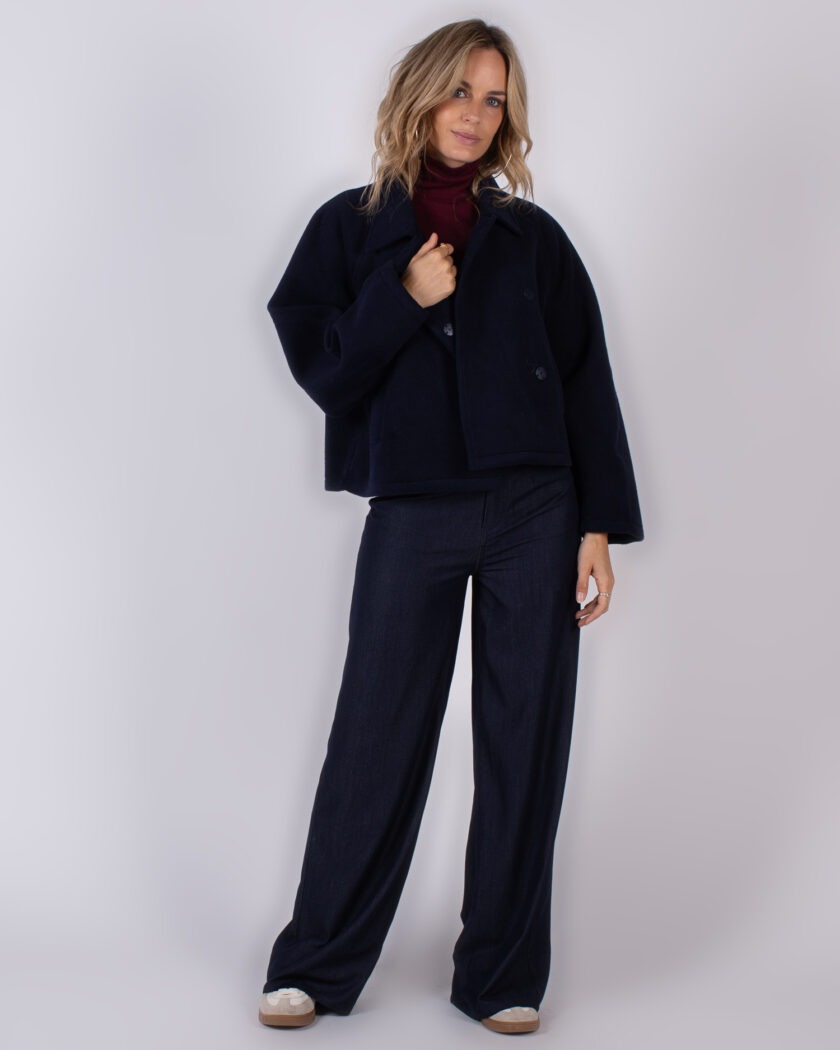 Vrouw poseert in een donkerblauwe wijde broek met bijpassende jas en een bordeauxrode coltrui uit de Studio AW25-26 collectie.