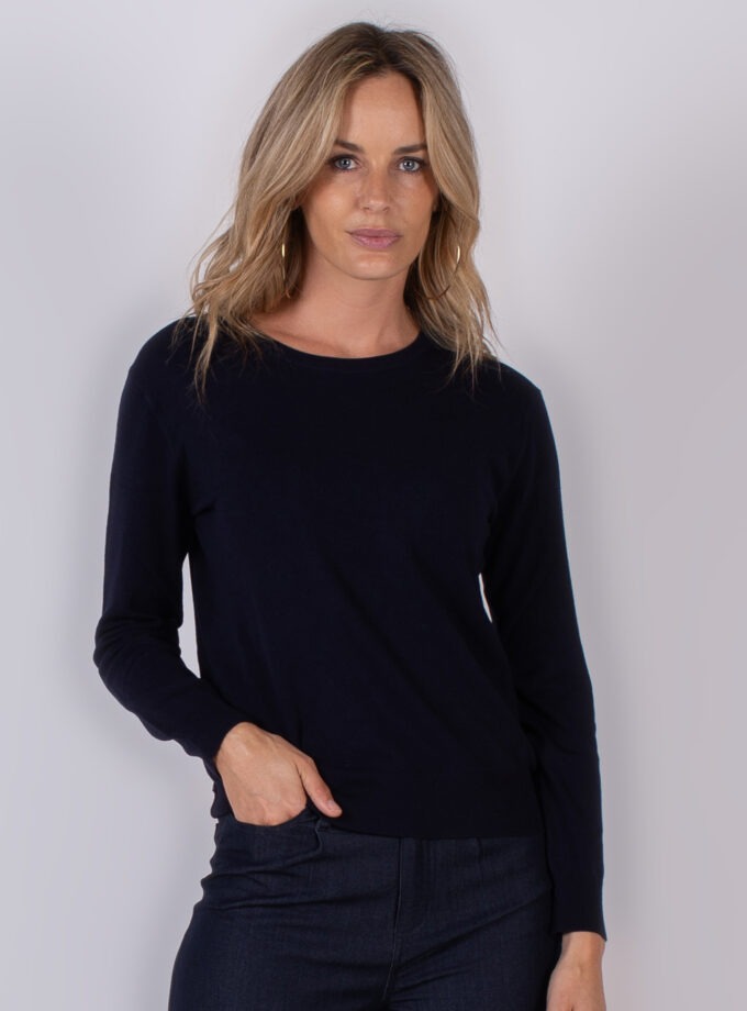 Vrouw met halflang blond haar draagt een donkerblauwe trui en donkere jeans tegen een neutrale achtergrond.