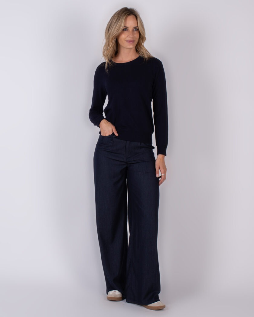 Vrouw draagt een donkerblauwe trui met wijde donkerblauwe broek en beige sneakers tegen een neutrale achtergrond.