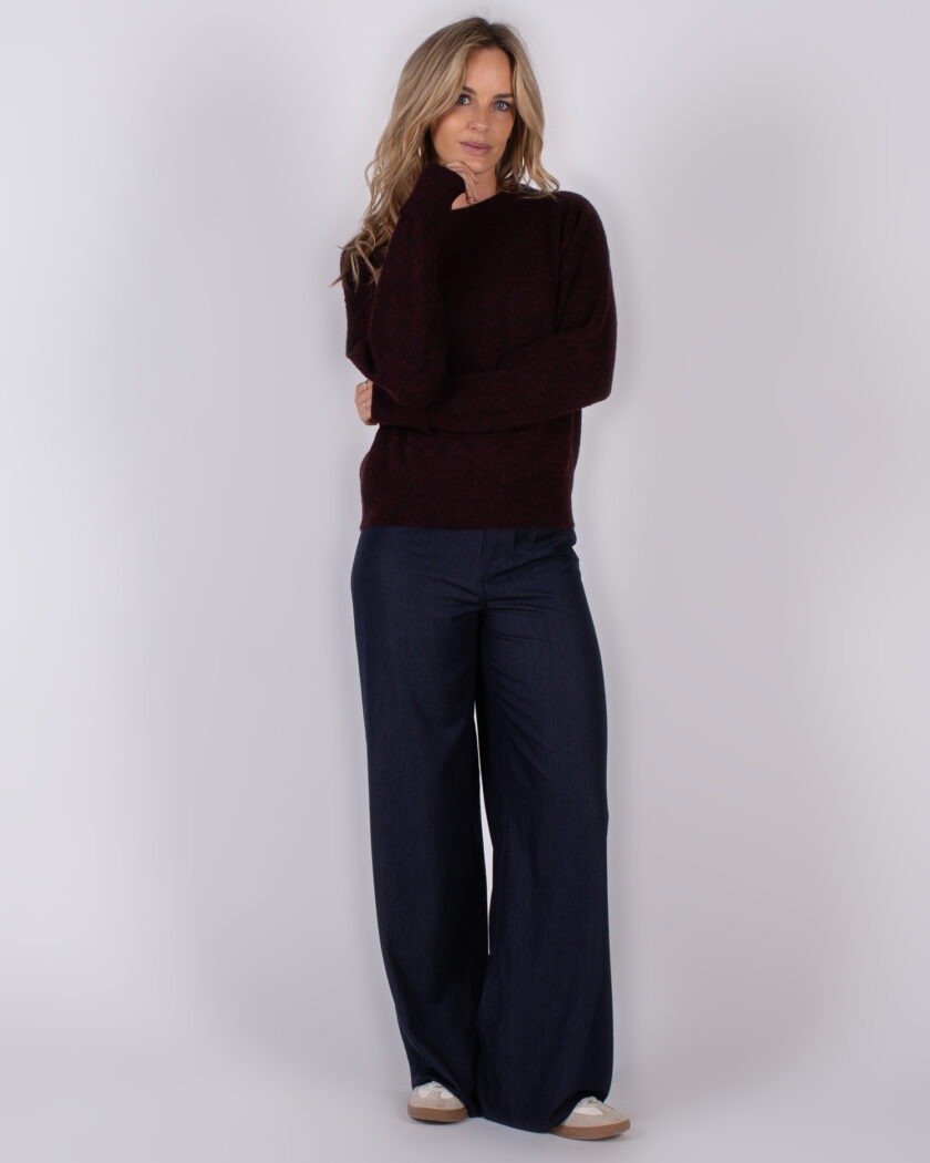 Vrouw draagt een donkerrode trui en wijde donkerblauwe broek uit de Studio AW25-26 collectie.