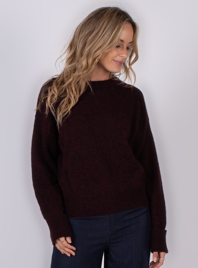 Vrouw draagt een donkerrode trui uit de Studio AW25-26 collectie en kijkt glimlachend omlaag.