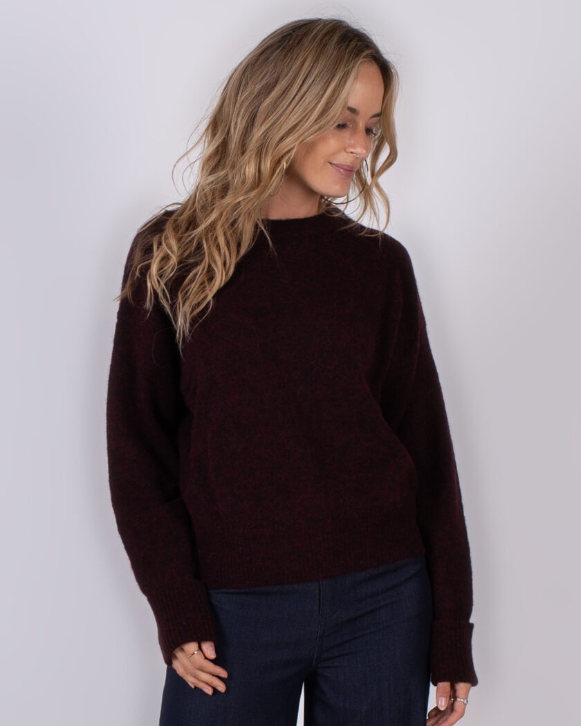Vrouw draagt een donkerrode trui uit de Studio AW25-26 collectie en kijkt glimlachend omlaag.