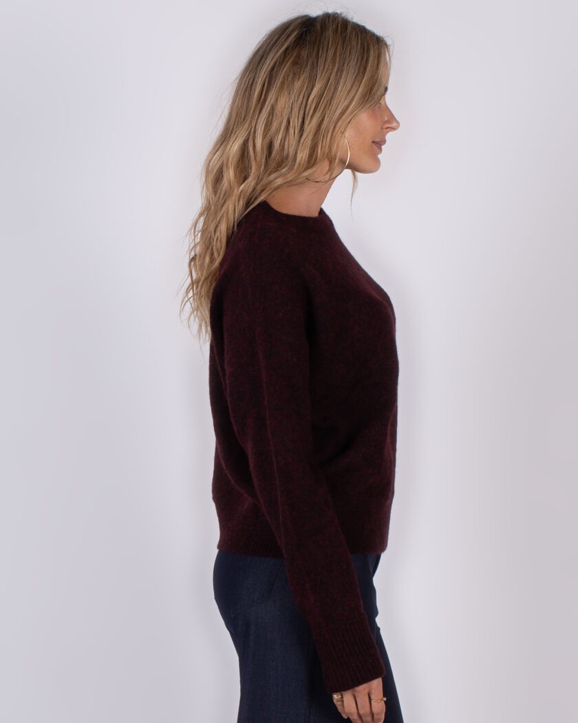 Vrouw in profiel draagt een donkerrode trui en donkere broek tegen een neutrale achtergrond.