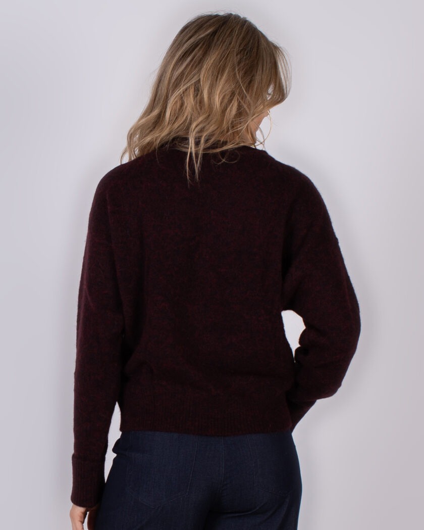 Vrouw van achteren in een donkerrode trui en donkere jeans, tegen een neutrale achtergrond.