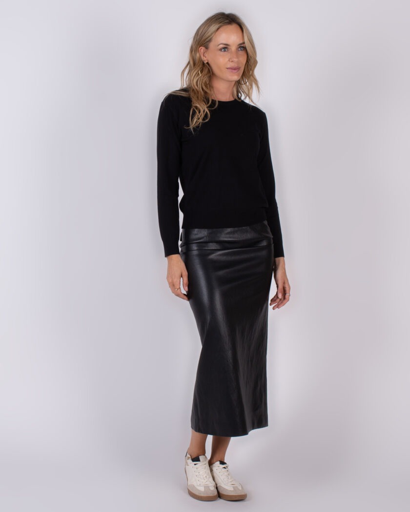 Vrouw in zwarte trui en lange zwarte leren rok met witte sneakers, gefotografeerd in studio voor Studio_AW25-26 collectie.