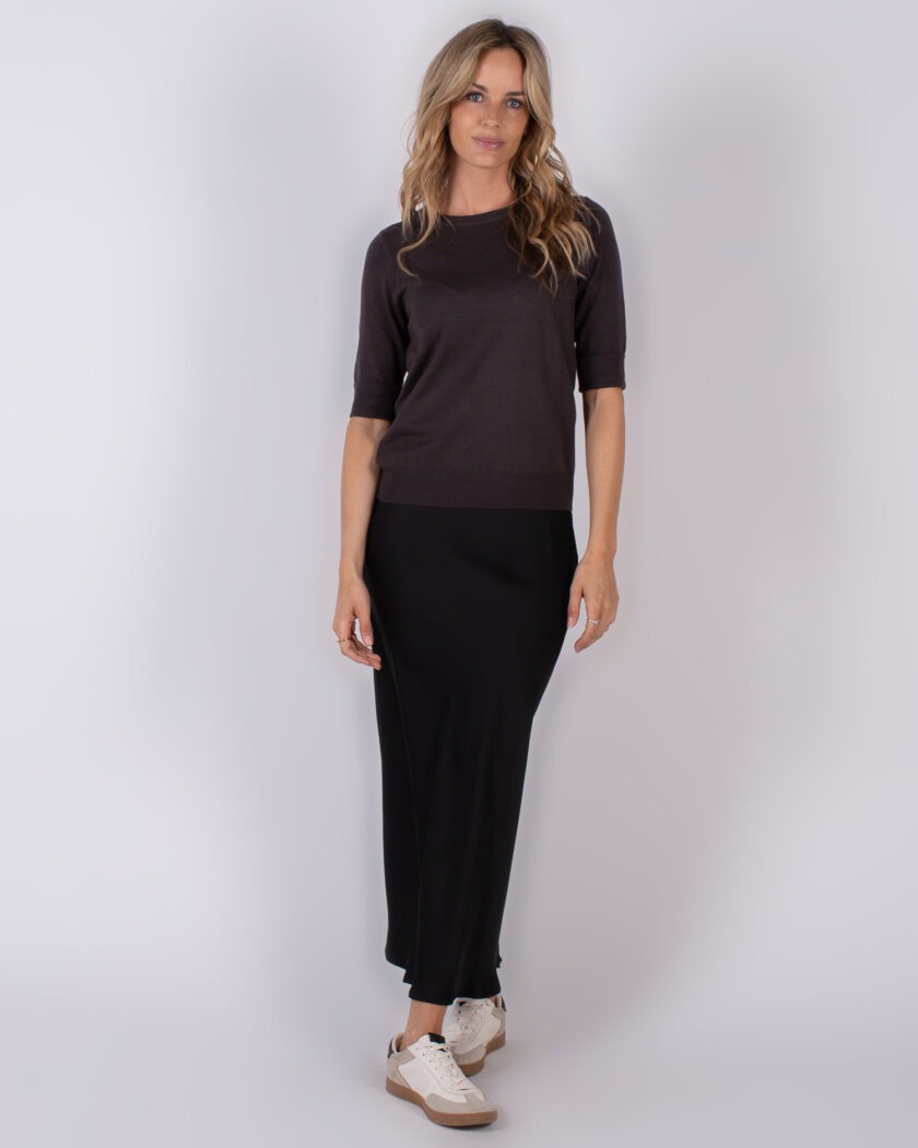Vrouw in zwarte top met halve mouw en zwarte lange rok, gecombineerd met witte sneakers, poseert tegen een neutrale achtergrond.
