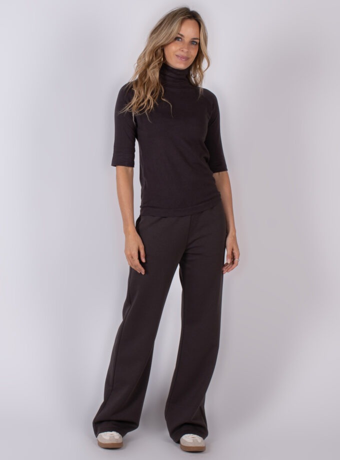 Vrouw poseert in een donkerbruin Studio AW25-26 outfit met hoge coltrui en wijde broek.