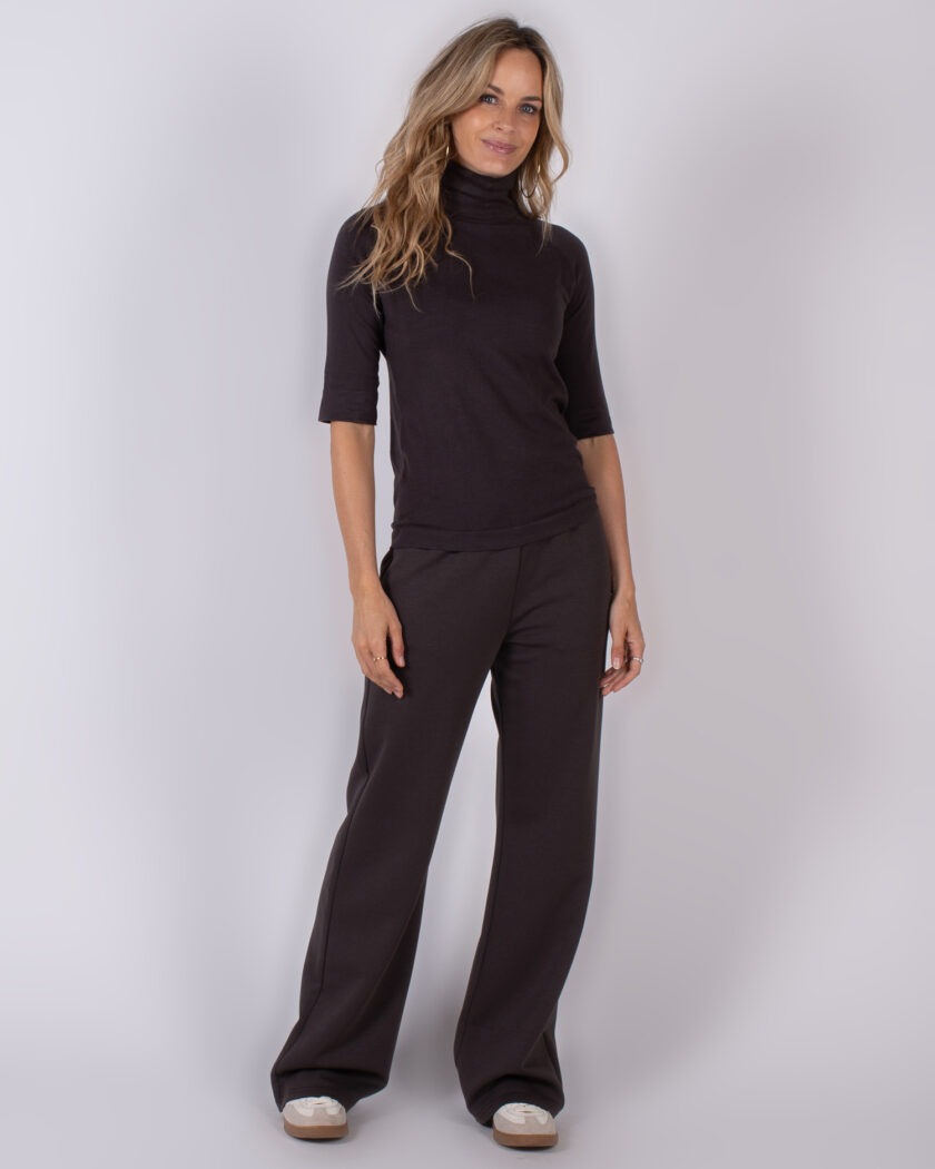 Vrouw poseert in een donkerbruin Studio AW25-26 outfit met hoge coltrui en wijde broek.
