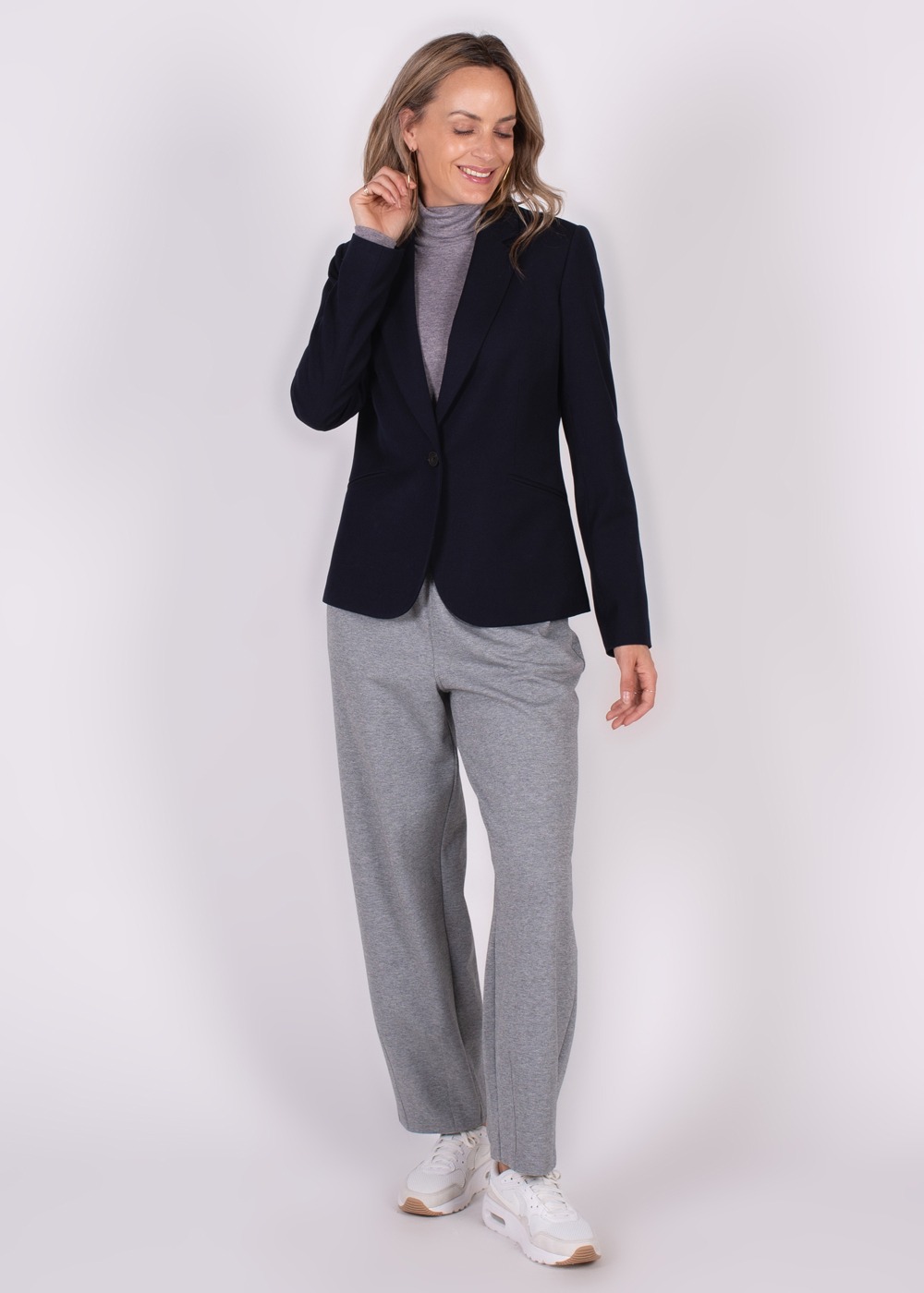 Een vrouw draagt een donkere Nikko Blazer gecombineerd met een grijze coltrui en grijze broek, terwijl ze glimlacht en haar rechterhand naar haar gezicht tilt.