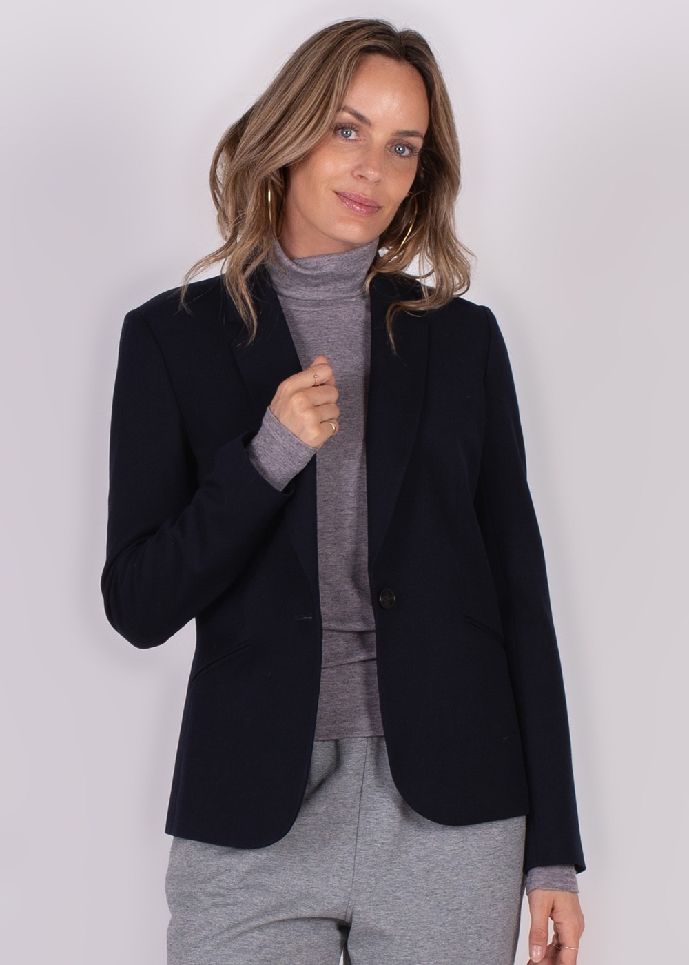Vrouwelijk model in een donkerblauwe blazer genaamd "Nikko" over een grijze coltrui, gefotografeerd tegen een effen achtergrond.