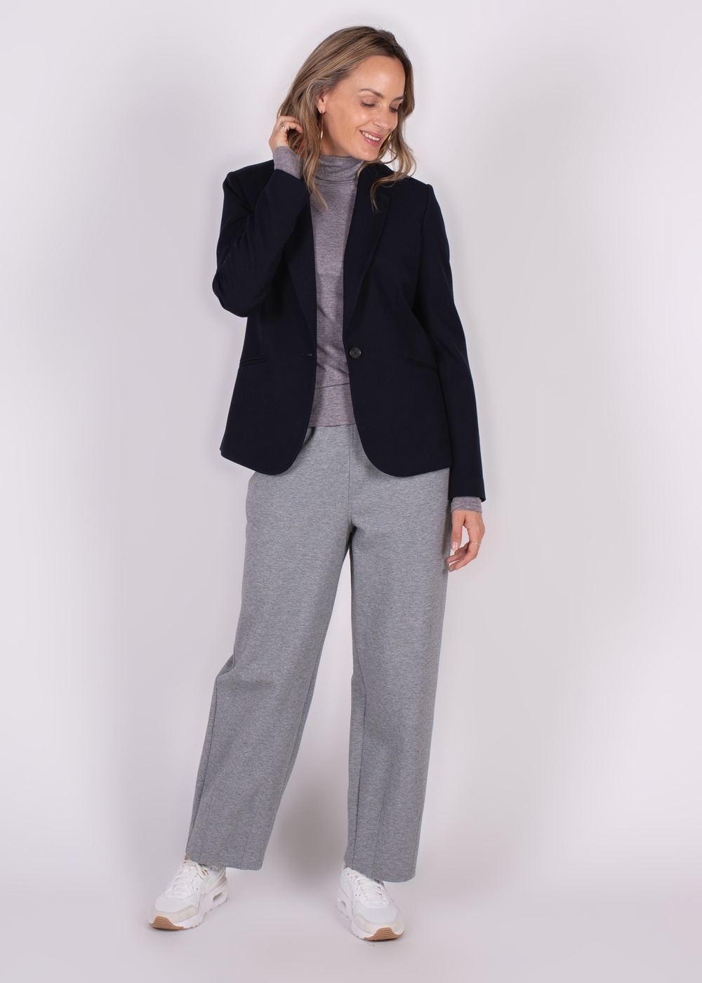 Een vrouw poseert in een donkerblauwe Nikko blazer, grijze coltrui, grijze broek en witte sneakers tegen een lichte achtergrond.