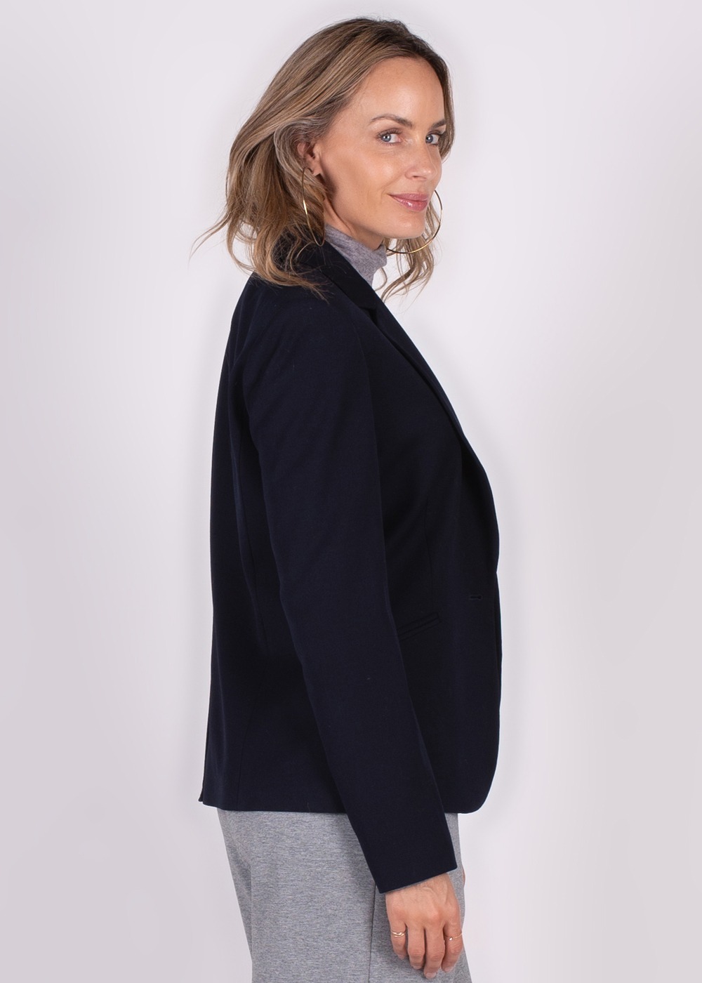 Een vrouw met losse haren poseert in zijaanzicht in een donkerblauwe Nikko blazer tegen een neutrale achtergrond.