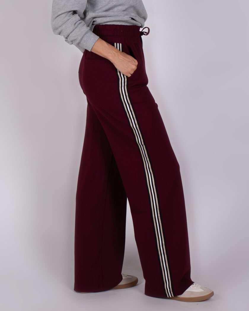 Bordeauxrode wide-leg broek met sportieve zijstrepen, gedragen met een grijze trui en witte sneakers.