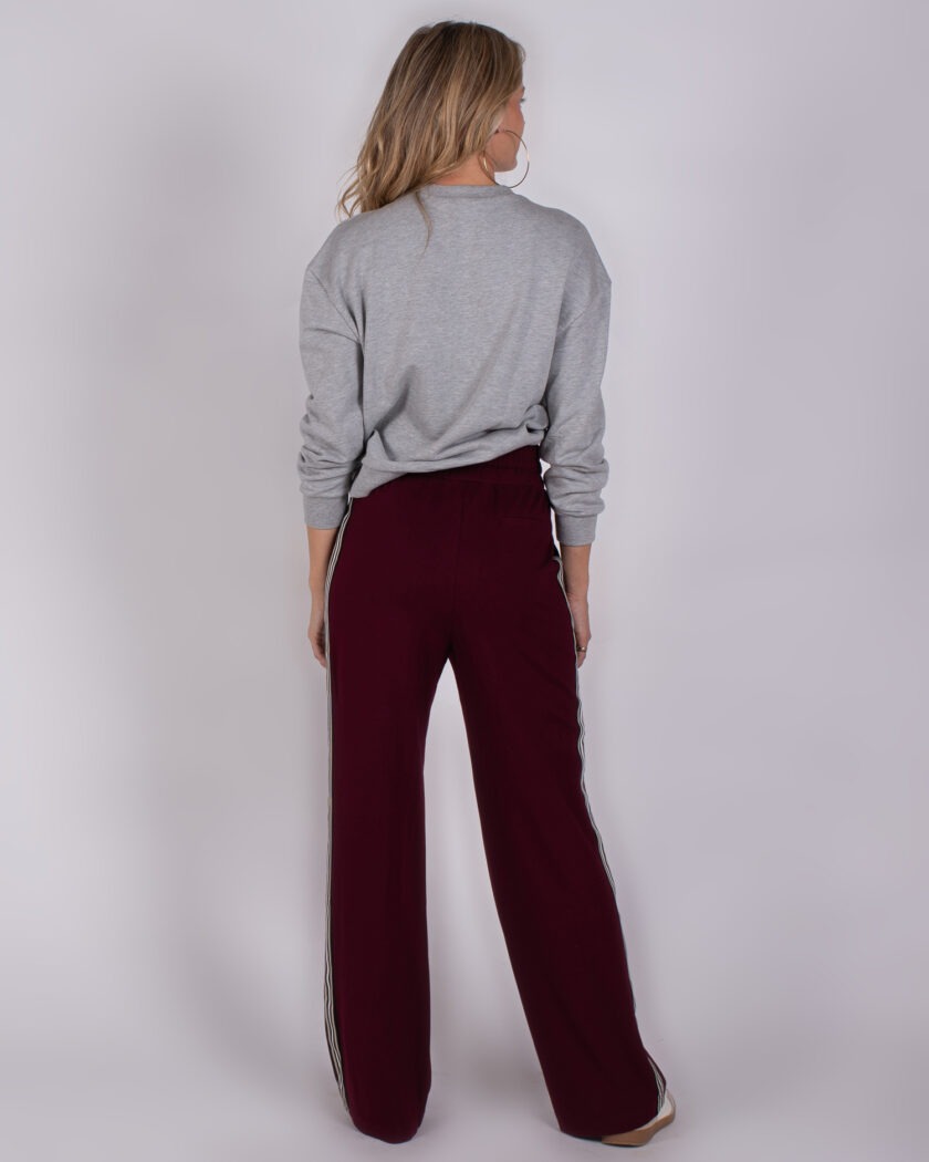 Vrouw van achteren in grijze trui en wijde bordeauxrode broek met sportieve zijstrepen.