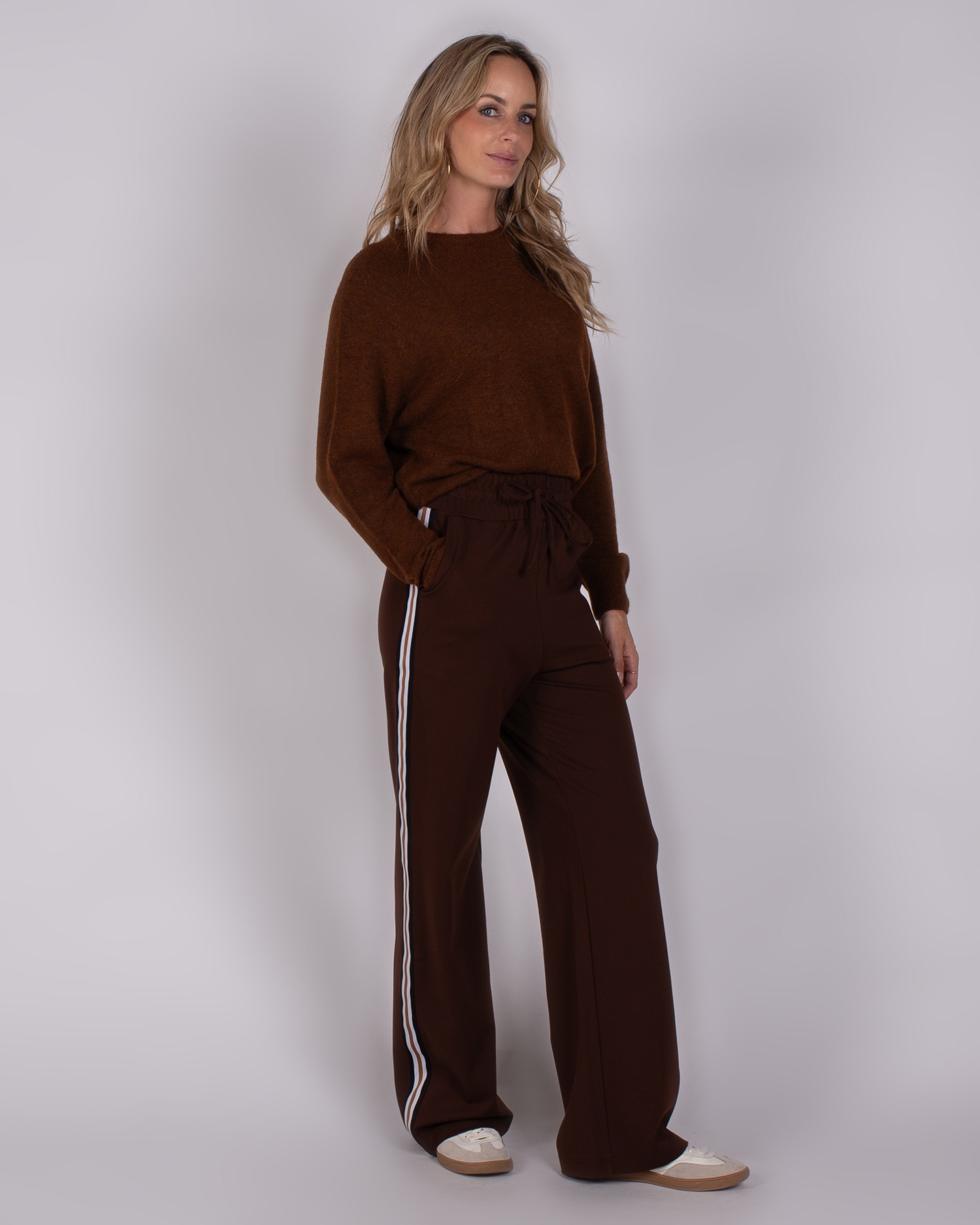 Vrouw draagt een bruine trui en wijde bruine broek met witte strepen in Studio_AW25-26-119.