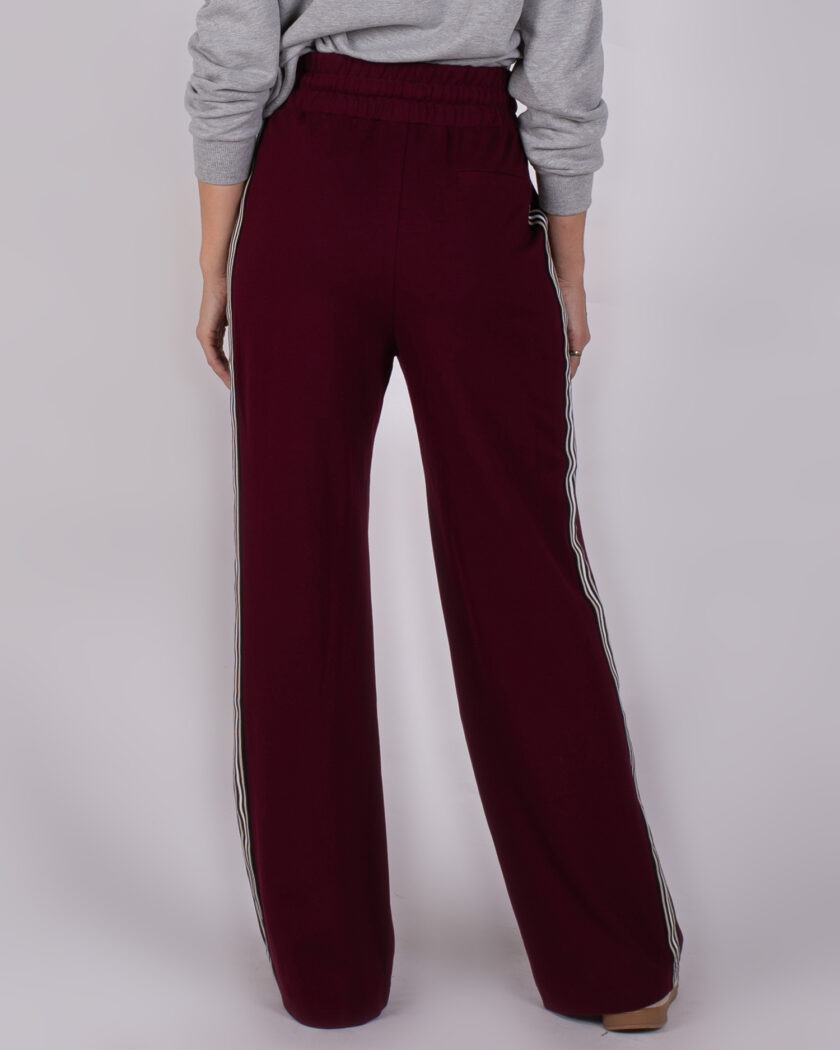 Achteraanzicht van een vrouw in wijde, bordeauxrode Studio AW25-26-12 broek met witte strepen langs de zijkanten.