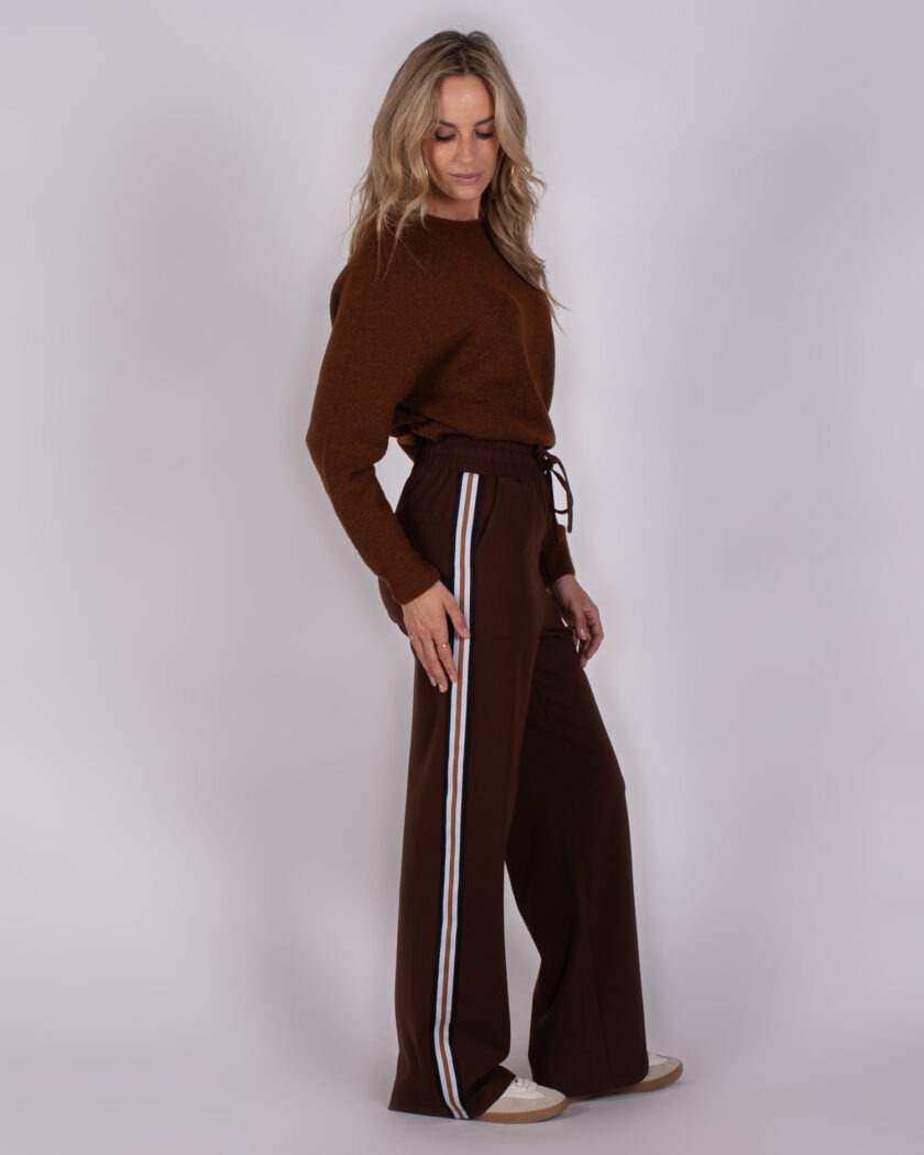 Vrouw draagt een bruine trui en wijde bruine broek met sportieve zijstrepen uit de Studio AW25-26 collectie.