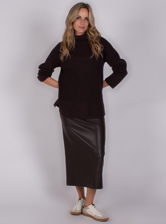 Vrouw in een Studio_AW25-26-281 outfit met een zwarte coltrui, zwarte leren midi-rok en witte sneakers.