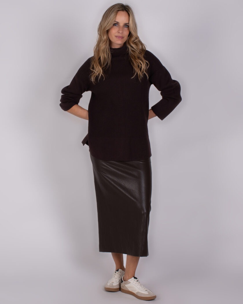 Vrouw in een Studio_AW25-26-281 outfit met een zwarte coltrui, zwarte leren midi-rok en witte sneakers.