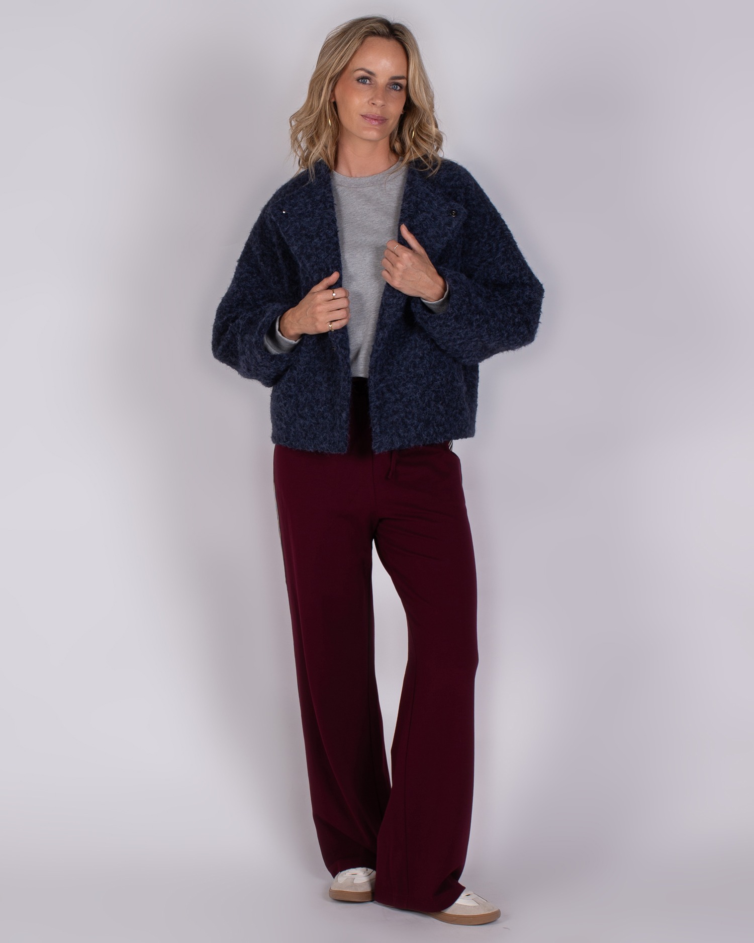 Studio_AW25-26-3 Vrouw in Studio AW25-26 outfit met een donkerblauwe jas, grijze trui en wijde bordeauxrode broek.