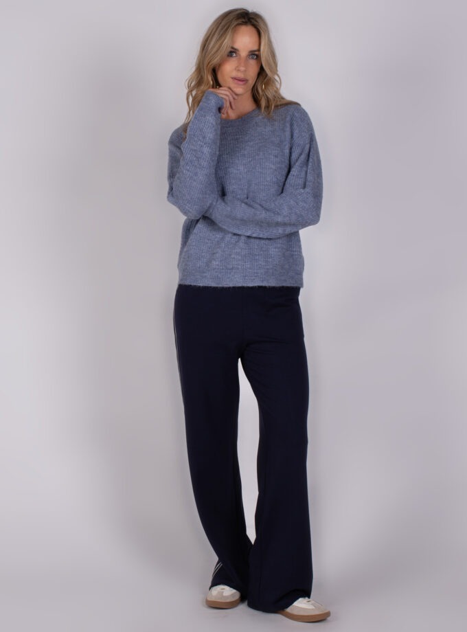 Vrouw draagt een lichtblauwe gebreide trui en donkerblauwe wijde broek met sneakers, uit de Studio AW25-26 collectie.