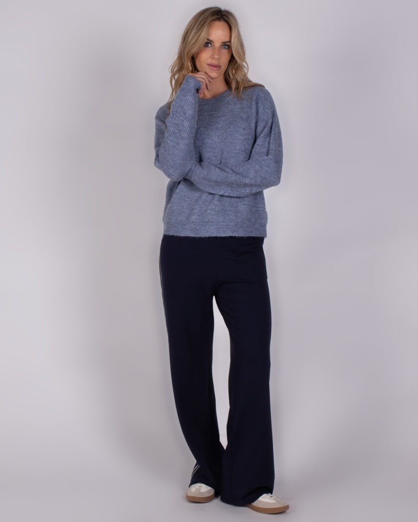 Vrouw draagt een lichtblauwe gebreide trui en donkerblauwe wijde broek met sneakers, uit de Studio AW25-26 collectie.
