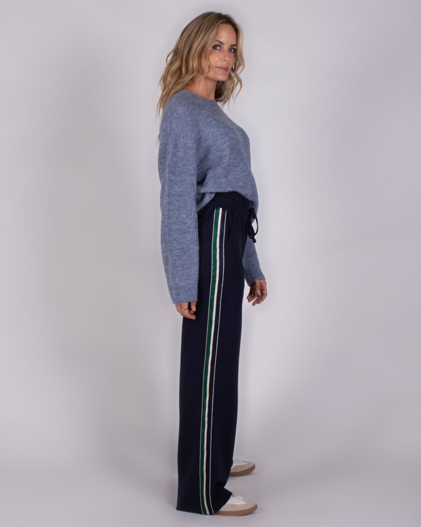 Vrouw in een lichtblauwe trui en donkerblauwe broek met gekleurde zijstrepen uit de Studio AW25-26 collectie.