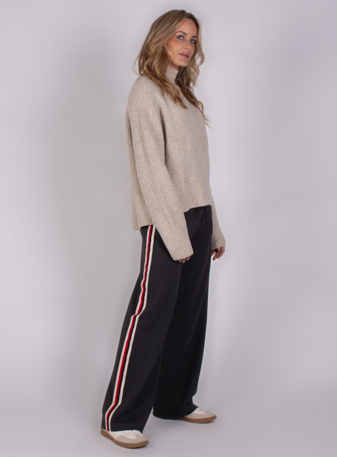 Vrouw in beige trui en zwarte broek met rode en witte zijstrepen uit de Studio AW25-26 collectie.