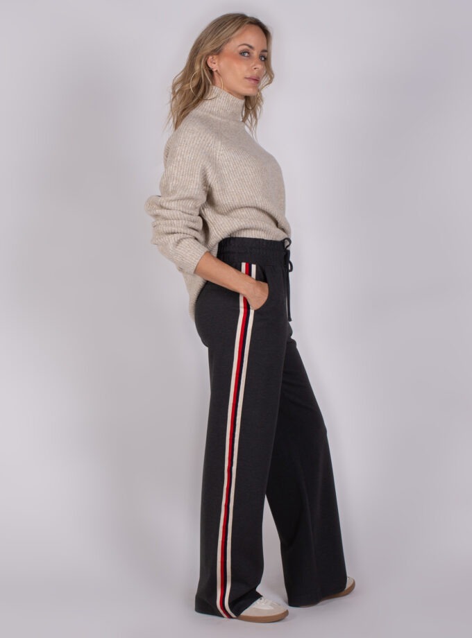 Vrouw in beige coltrui en donkergrijze broek met rood-wit gestreepte zijstreep, kijkend opzij.