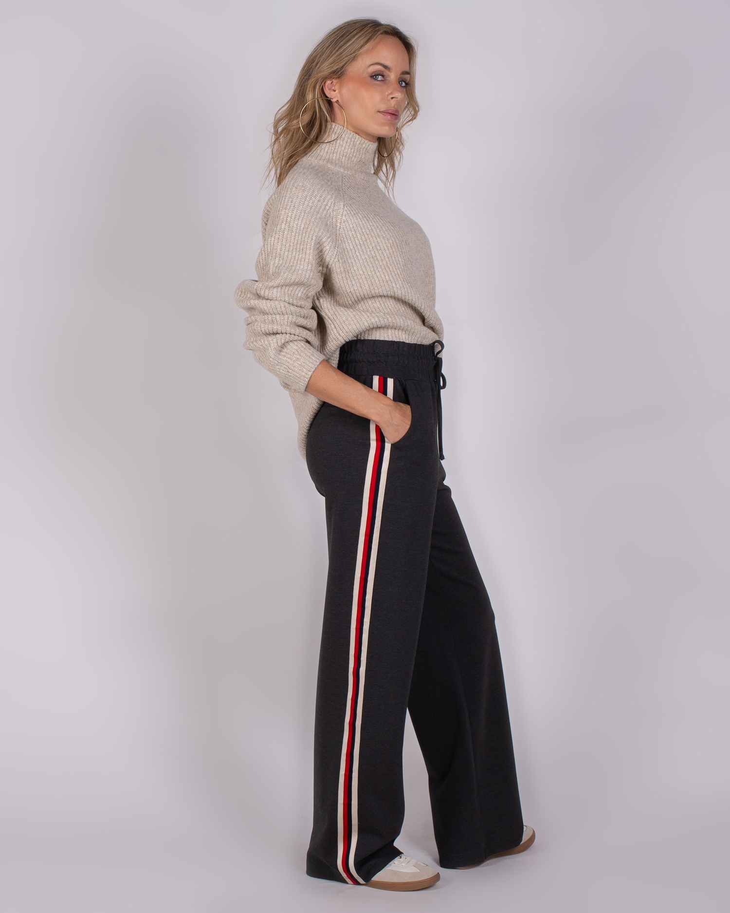 Vrouw in beige coltrui en donkergrijze broek met rood-wit gestreepte zijstreep, kijkend opzij.