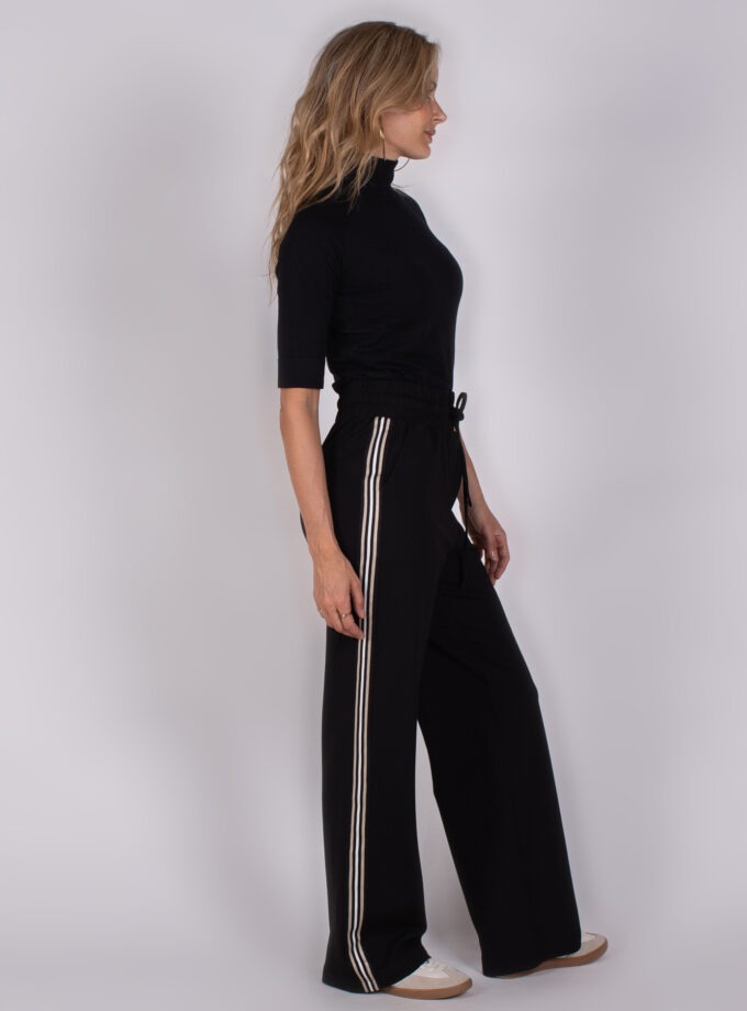 Vrouw in zwarte coltop en wijde zwarte broek met witte strepen aan de zijkant, staand in zijaanzicht.