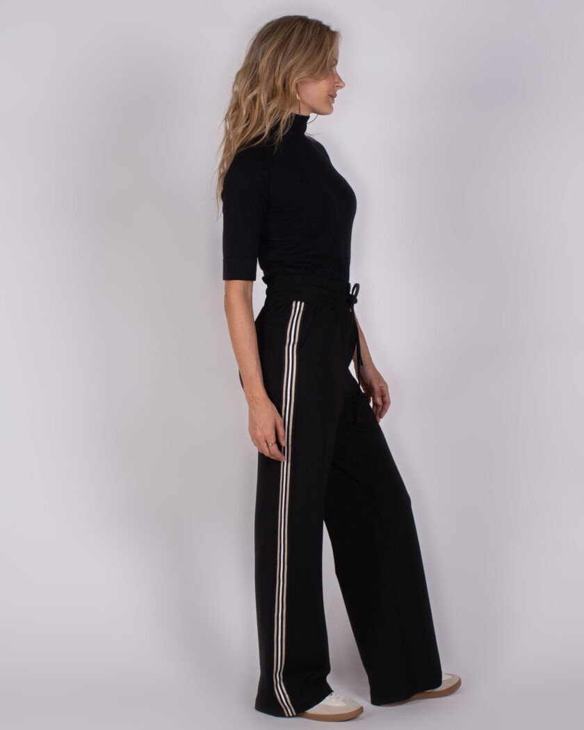 Vrouw in zwarte coltop en wijde zwarte broek met witte strepen aan de zijkant, staand in zijaanzicht.