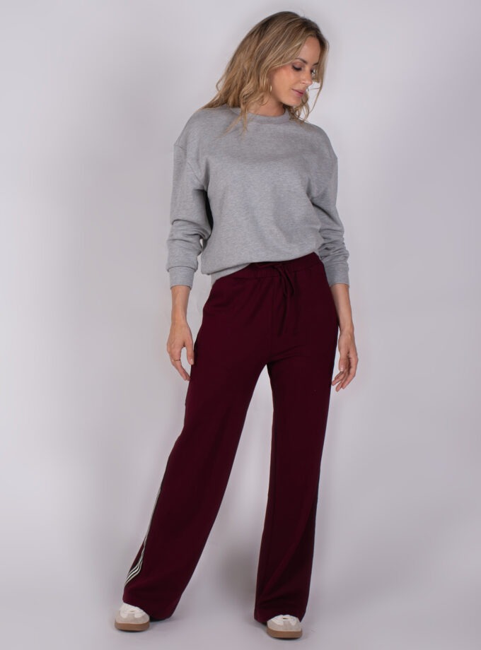 Vrouw in grijze trui en bordeauxrode wijde broek met sportieve sneakers uit de Studio AW25-26 collectie.