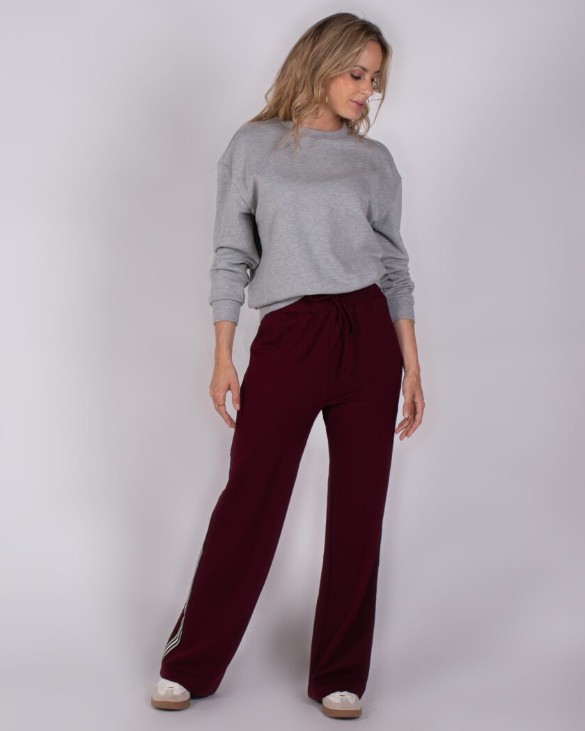 Vrouw in grijze trui en bordeauxrode wijde broek met sportieve sneakers uit de Studio AW25-26 collectie.