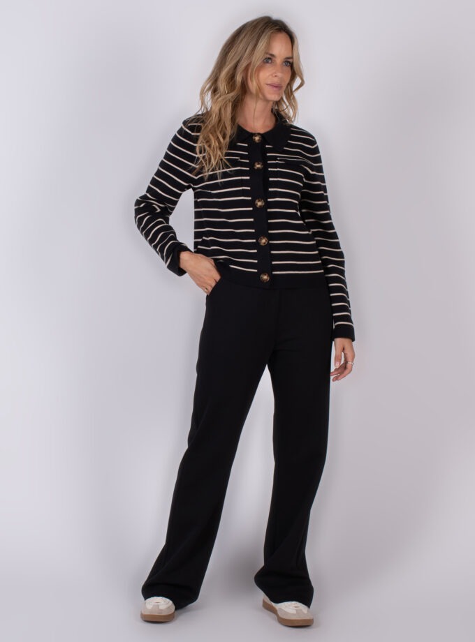 Vrouw poseert in een zwarte outfit met beige strepen en grote knopen uit de Studio AW25-26 collectie.