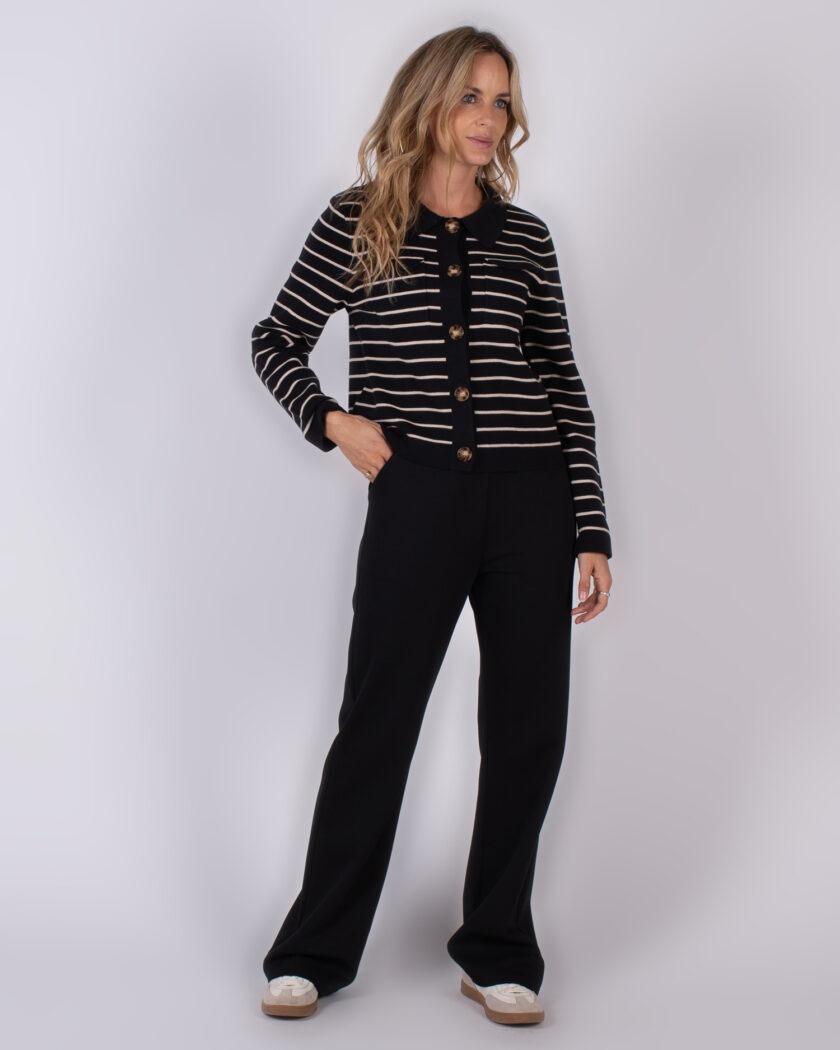 Vrouw poseert in een zwarte outfit met beige strepen en grote knopen uit de Studio AW25-26 collectie.