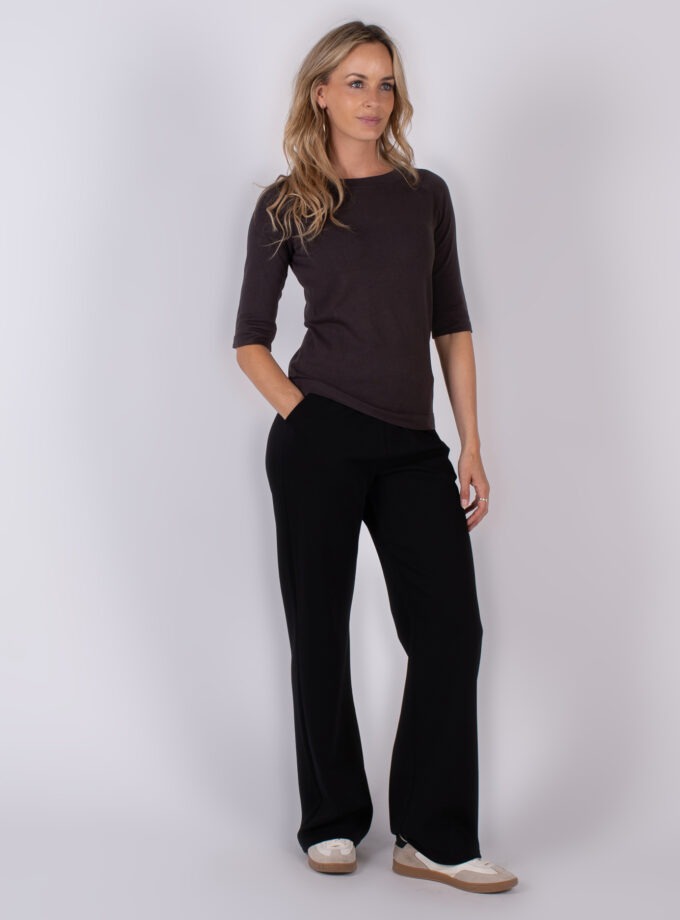 Vrouw poseert in een donkerbruine top met driekwartsmouwen en zwarte wijde broek uit de Studio AW25-26 collectie.