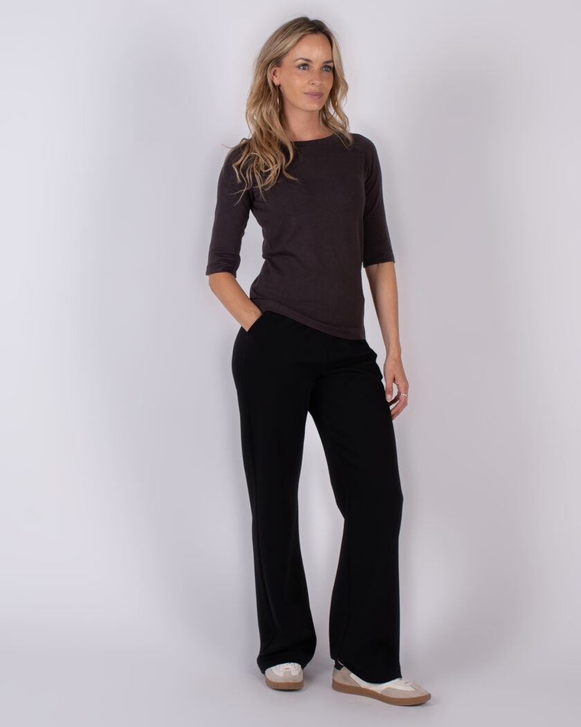 Vrouw poseert in een donkerbruine top met driekwartsmouwen en zwarte wijde broek uit de Studio AW25-26 collectie.