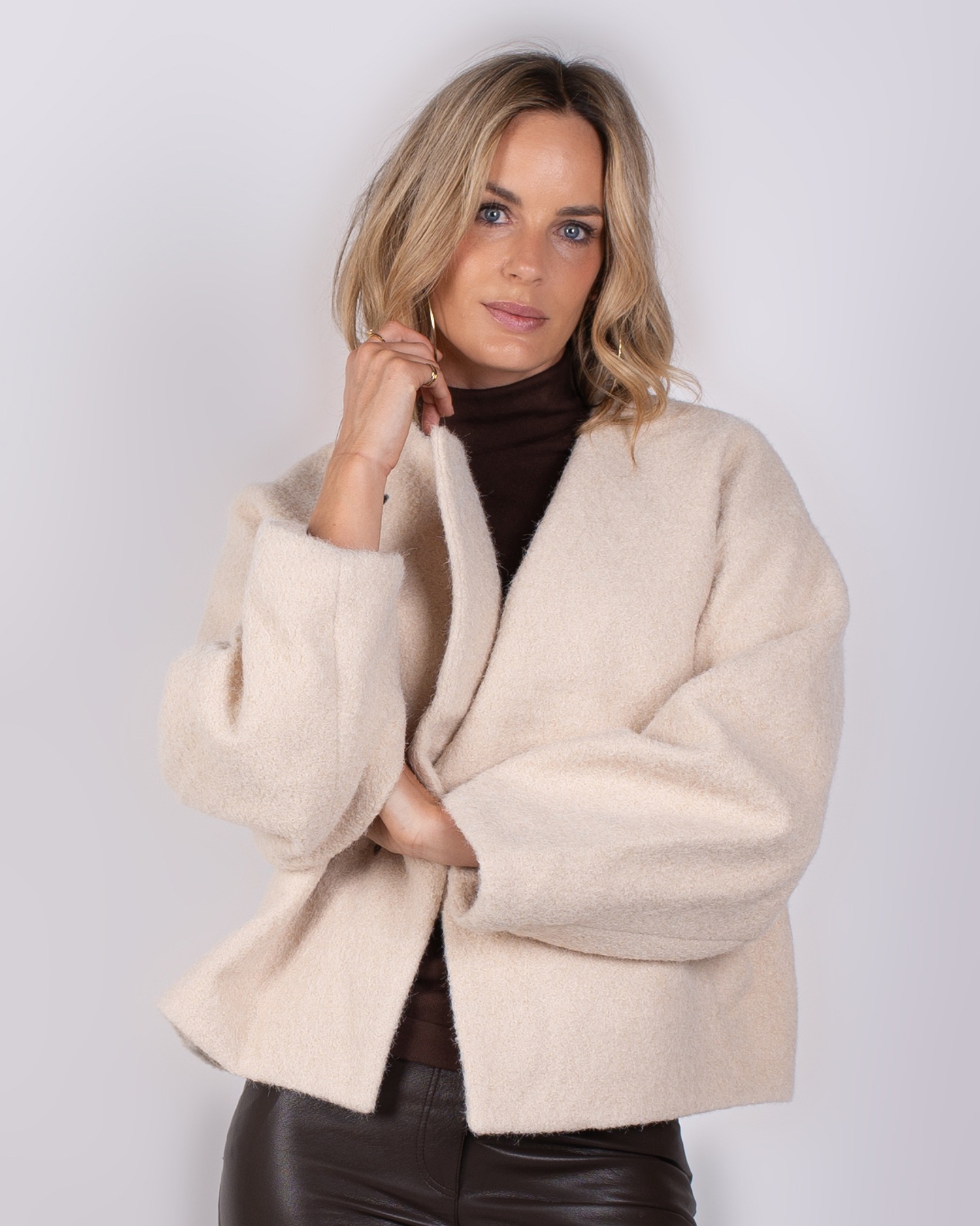 Studio_AW25-26#3-13 Vrouw draagt een beige, wollen jas van Studio AW25-26#3-13 over een donkerbruine coltrui.