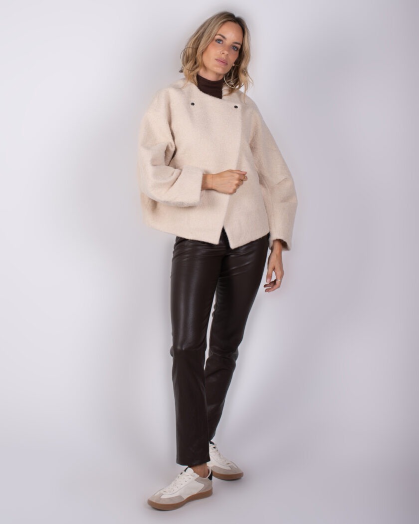 Vrouw draagt een beige oversized jas, donkerbruine leren broek en witte sneakers tegen een neutrale achtergrond.